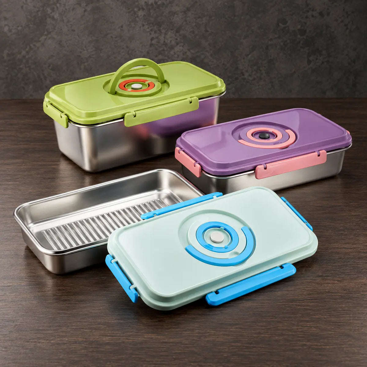 Three rectangular stainless steel food storage containers with green, purple, and light-blue latching lids, one shown open