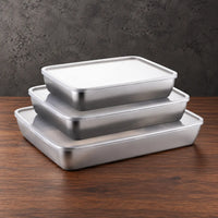 Three wide rectangular stainless steel food storage containers stacked with light-colored lids